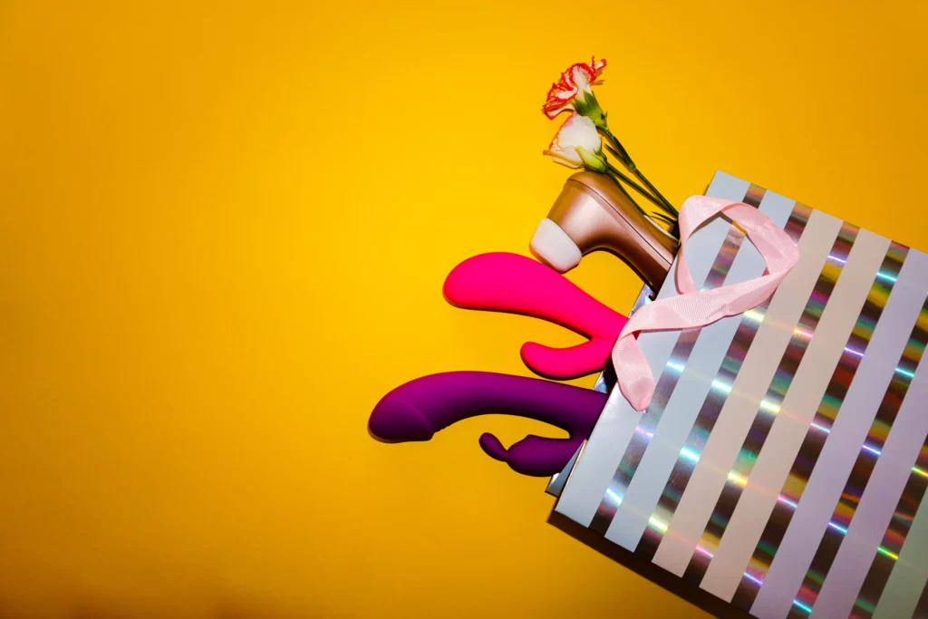 Several colorful sex toys arranged inside and around a striped shopping bag on a yellow background