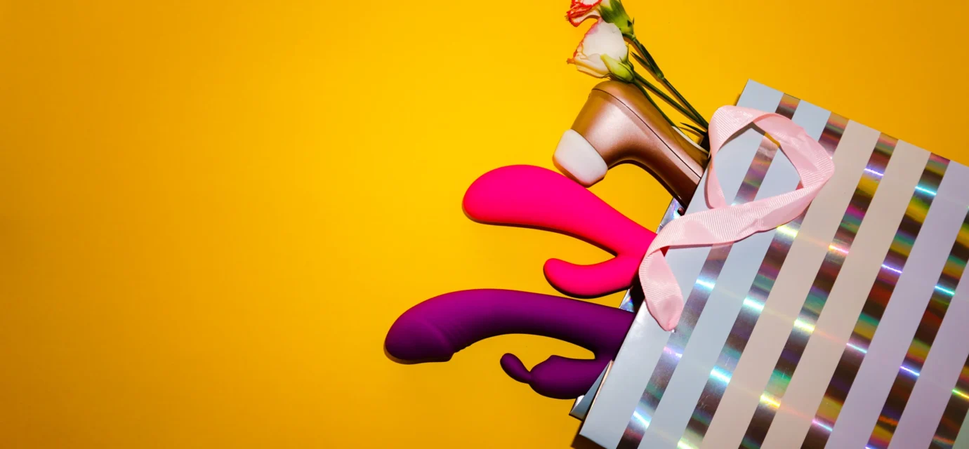 Several colorful sex toys arranged inside and around a striped shopping bag on a yellow background