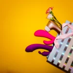 Several colorful sex toys arranged inside and around a striped shopping bag on a yellow background