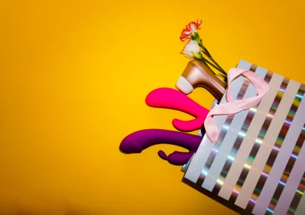 Several colorful sex toys arranged inside and around a striped shopping bag on a yellow background