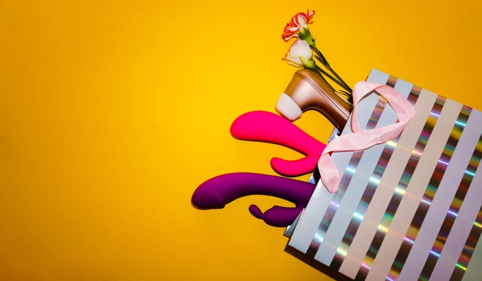 Several colorful sex toys arranged inside and around a striped shopping bag on a yellow background